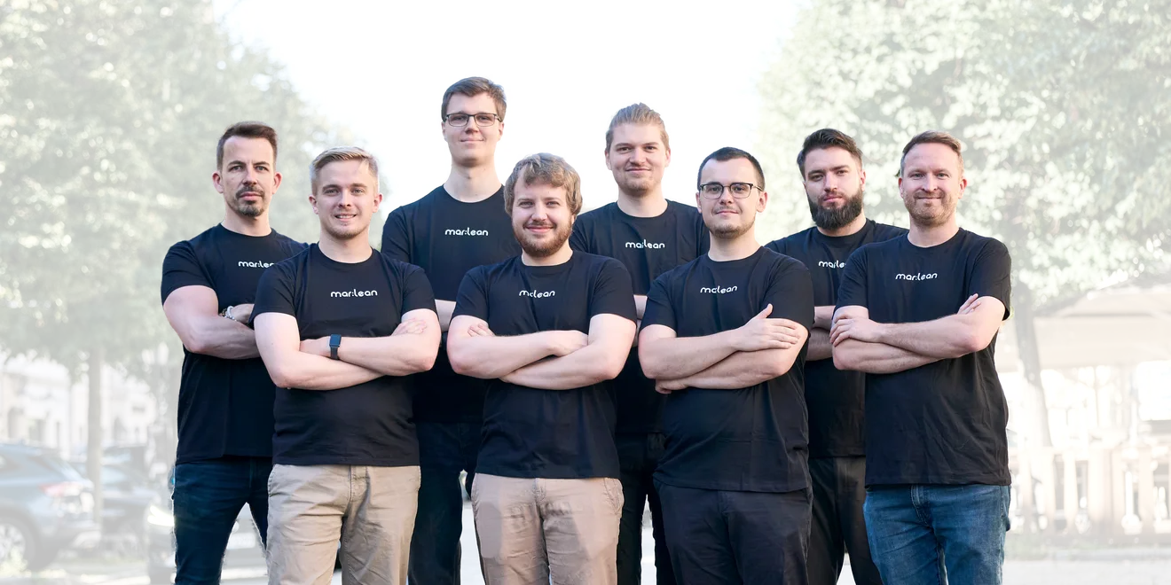 Eight members of the mar:lean team in black mar:lean T-shirts, arms crossed, on a street in daylight